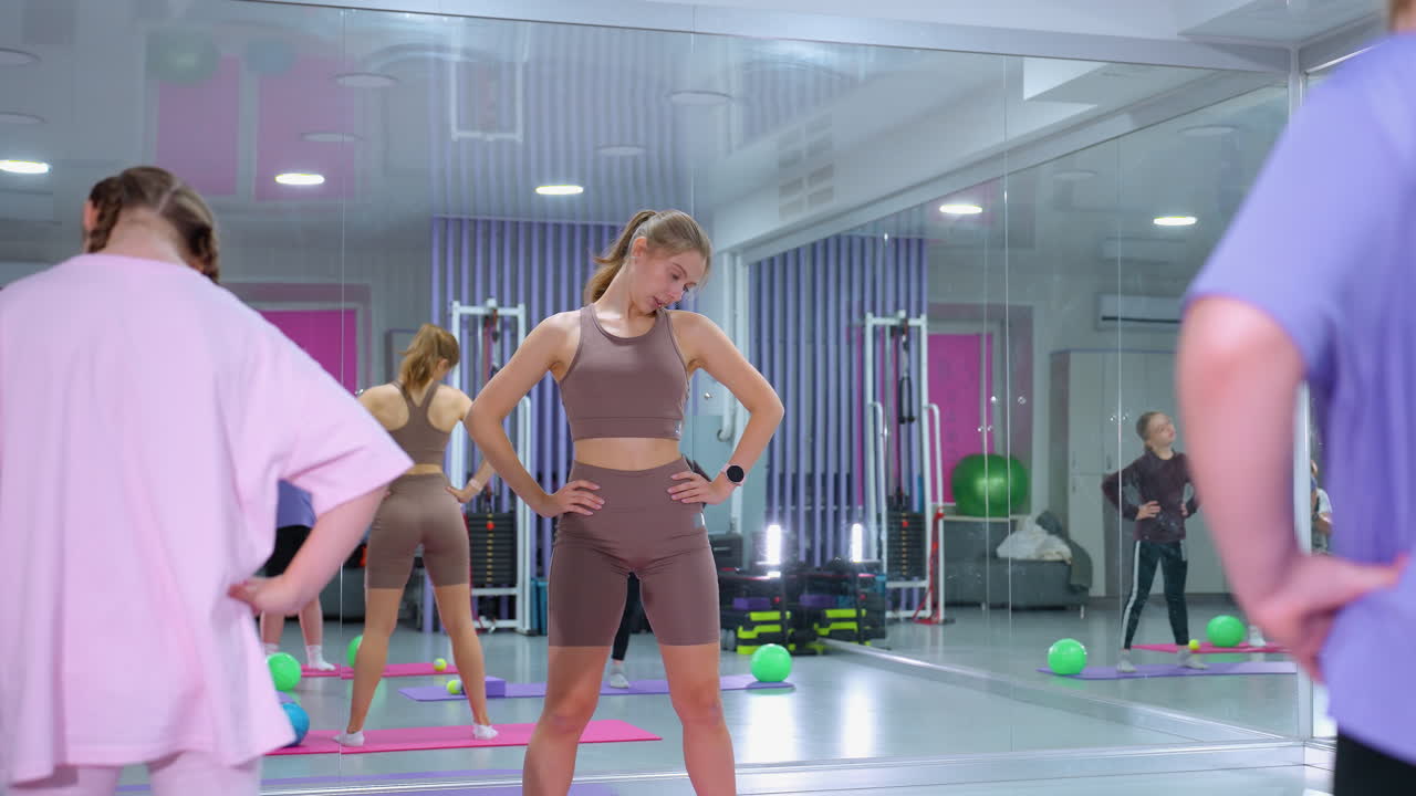 체육관 강사는 두 명의 여성이 목을 회전하는 운동을 이끌고, 밝은 피트니스 스튜디오에서 허리에 손을 고, 큰 거울은 다른 참가자를 검은색으로 반영합니다.