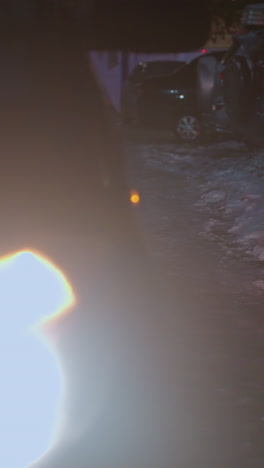 faros de automóviles iluminados proyectando un haz brillante en una carretera helada rodeada de nieve y vegetación dispersa, con coches estacionados borrosos y edificios débiles en el fondo