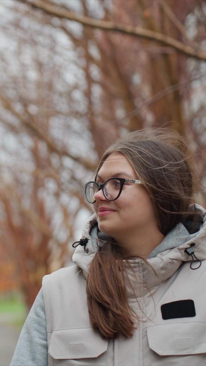 Una persona indossa occhiali neri e una giacca chiara sopra un top grigio. I capelli sciolti del soggetto le cadono sulla spalla mentre guarda. L'ambiente esterno include alberi spogli e luce soffusa.