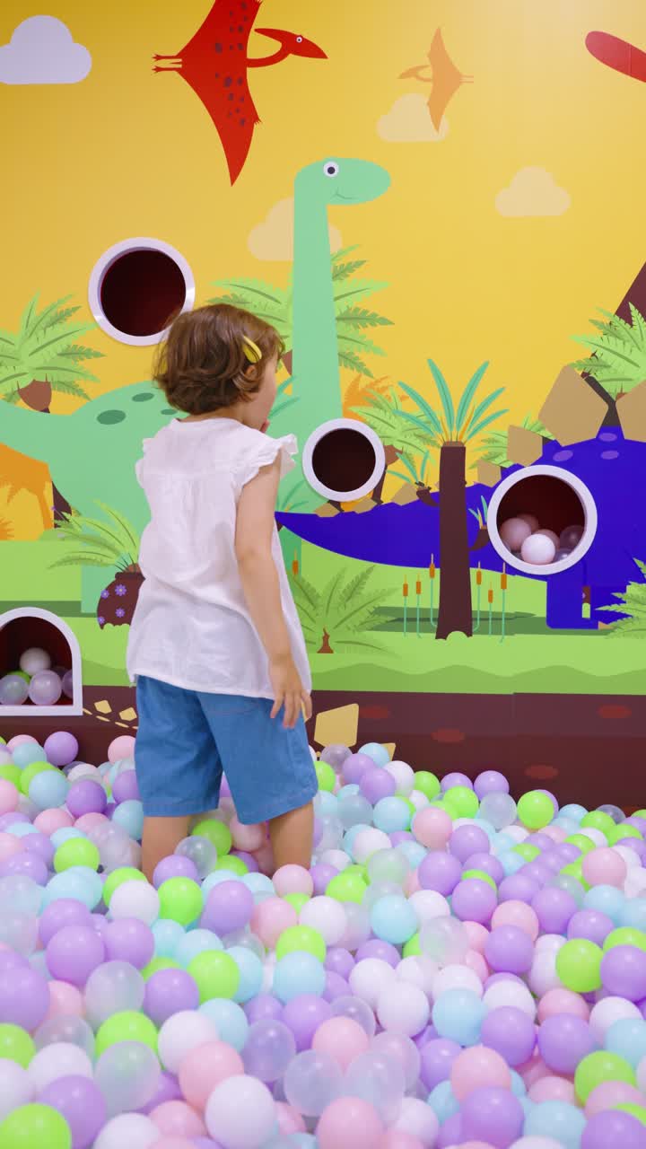 Five-year-old child plays in a colorful indoor playground ball pit, throwing balls into wall holes decorated with dinosaurs and volcanoes