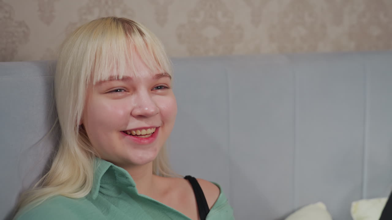 Two young women lie on bed, smiling and laughing in joyful conversation, one wears mint green shirt, other wears black pajamas, soft pillows and warm lighting add to cozy indoor relaxation scene