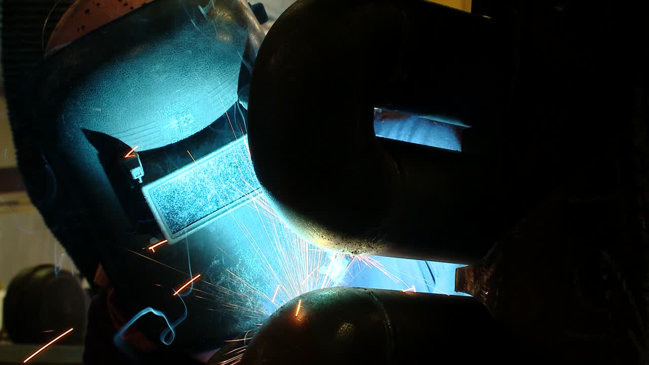 Reverse close-up zooming out shot of a welder in full protective HSE kit welding pipes at a factory with intense light and nice sparks from the welding operation
