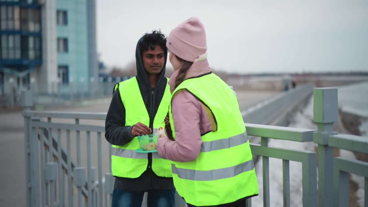 Man in hoodie and neon safety vest holding container of food talking with woman in pink jacket and matching neon vest on bridge near river during cold overcast day, showing concept of outdoor work
