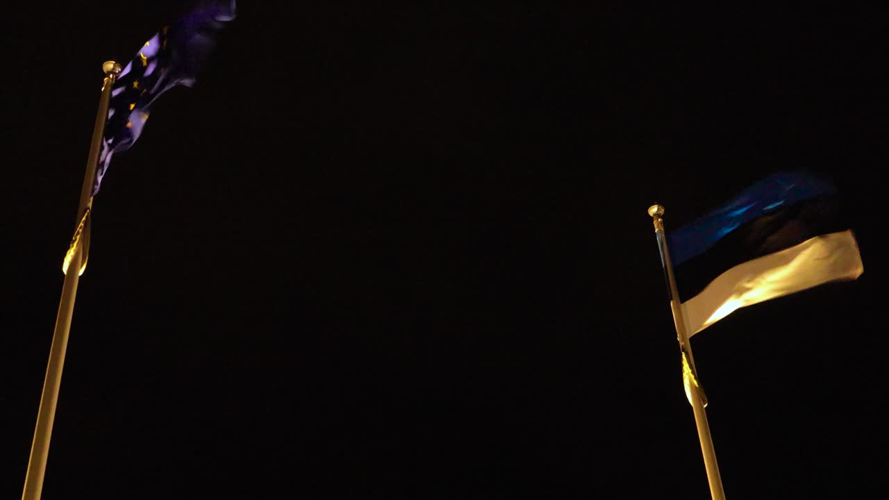 Duas bandeiras da União Europeia e da Estônia tremulando ao vento em mastros dourados contra um céu escuro. Iluminadas por luzes brilhantes vindas de baixo. Simbolizando o orgulho nacional e as relações internacionais