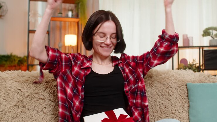 Giovane donna felice che apre un regalo a casa, sorpresa e gioiosa, reagisce con un ampio sorriso.
