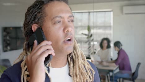 busy biracial male creative talking on smartphone and colleagues talking in background, slow motion