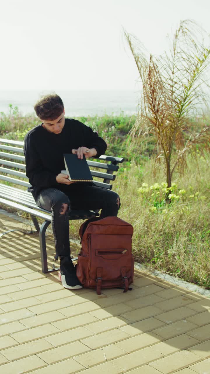 Young Man On A Bench Near The Sea Picking A Notebook From The Backpack