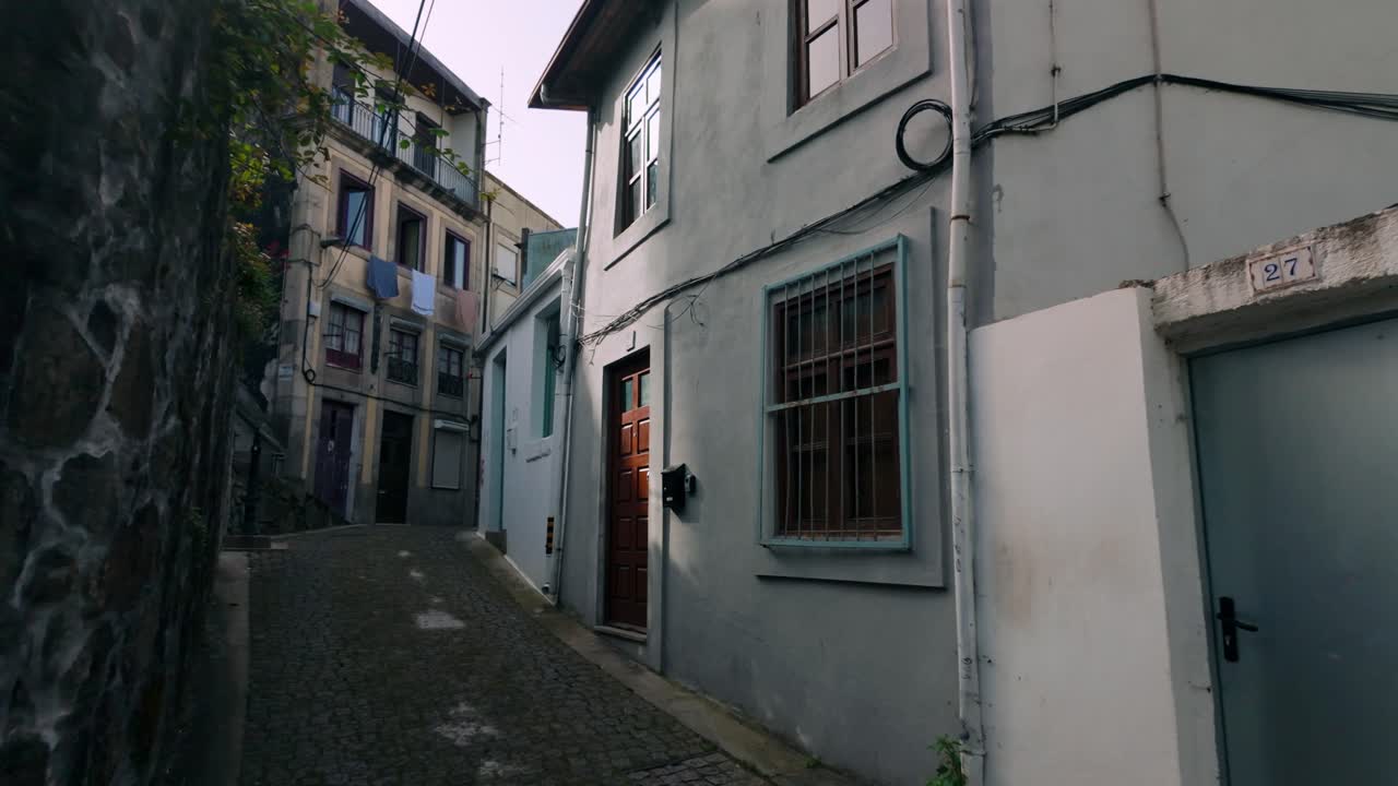 Stretto vicolo acciottolato in un'incantevole città europea, fiancheggiato da edifici storici e sbiaditi. Un vecchio muro di pietra sulla sinistra offre texture e un senso di età e tradizione.