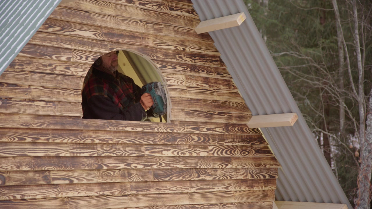 Manitas usando sierra de calar para cortar una ventana arqueada en la pared de madera de una minicasa.
