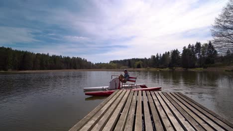 young man on a pedal boat leaving a wooden pier on a pond surrounded by a wood