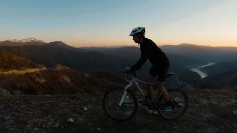 Man-riding-a-bike-at-the-top-of-a-mountain-with-beautiful-canyon-lake-at-sunset-in-the-background