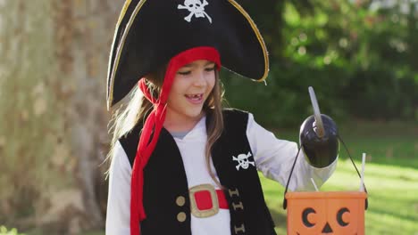 Video-of-happy-caucasian-girl-in-pirate-costume-holding-halloween-trick-or-treat-basket-in-garden