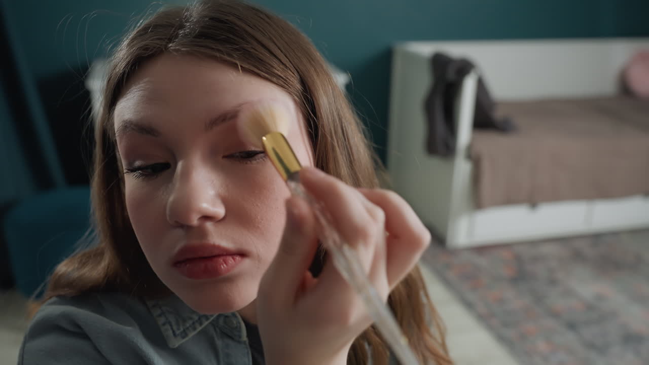 Creatore caucasico applica il fard per un tutorial, inquadrature di profilo passo-passo, pennello soffice che sfuma lungo la guancia, parete verde acqua e letto sullo sfondo, espressione concentrata, energia da influencer