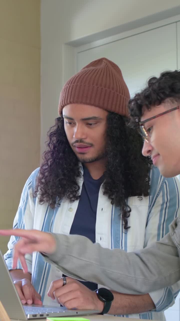 Vertical video: Typing beanie-wearing man on laptop at desk, peer joining to help and pointing