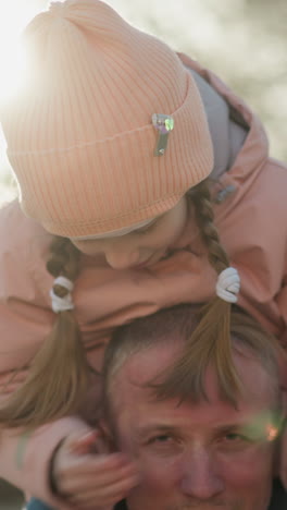 dulce momento en el parque como una niña con una gorra rosa cubre los ojos de su padre mientras se sienta en sus hombros, ambos disfrutando de la interacción lúdica