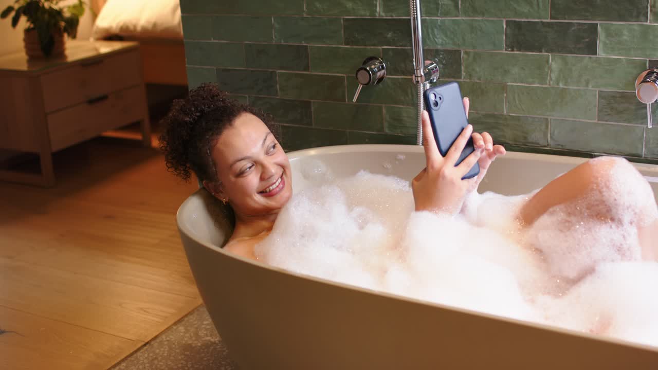 African American woman receiving text, reclining deeper in bathtub, holding smartphone, laughing