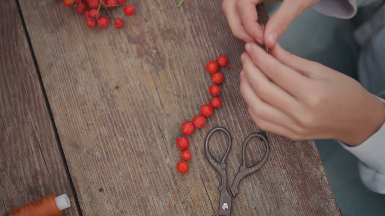 Primo piano di mani che infilano bacche rosse su un tavolo di legno che mostrano mani femminili che creano una ghirlanda stagionale con rocchetto e forbici, texture rustica, atmosfera autunnale calma, focus su grappoli di sorbo perlati e stabilità.