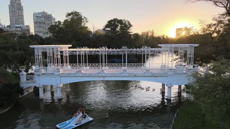 Statische-Aufnahme-Von-Touristen,-Die-Mit-Dem-Tretboot-über-Die-Brücke-Paddeln,-Gefolgt-Von-Einem-Schwarm-Wasservögel-An-Einem-Ruhigen-Nachmittag-Mit-Einem-Großen,-Hellen-Sonnenuntergang-Im-Hintergrund-Im-Rosengarten-Parque-Tres-De-Febrero