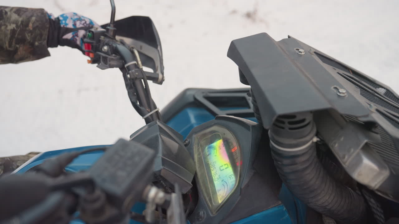 Manubrio e cruscotto di un ATV nella neve, mano guantata gira l'acceleratore e avvia l'accensione, il quadro strumenti si illumina, il pilota si prepara per un giro fuoristrada, portapacchi anteriore e telaio dello specchietto in un freddo paesaggio invernale.
