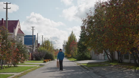 young boy in blue hoodie and black beanie walks down residential street with paper bag swaying in hand, surrounded by vibrant autumn foliage, colorful houses, and cloudy skies