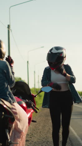dos mujeres con cascos están de pie al lado de la carretera, con una sentada en una motocicleta mientras la otra camina hacia ella, en el fondo, los coches se acercan, y un motociclista saluda mientras pasan