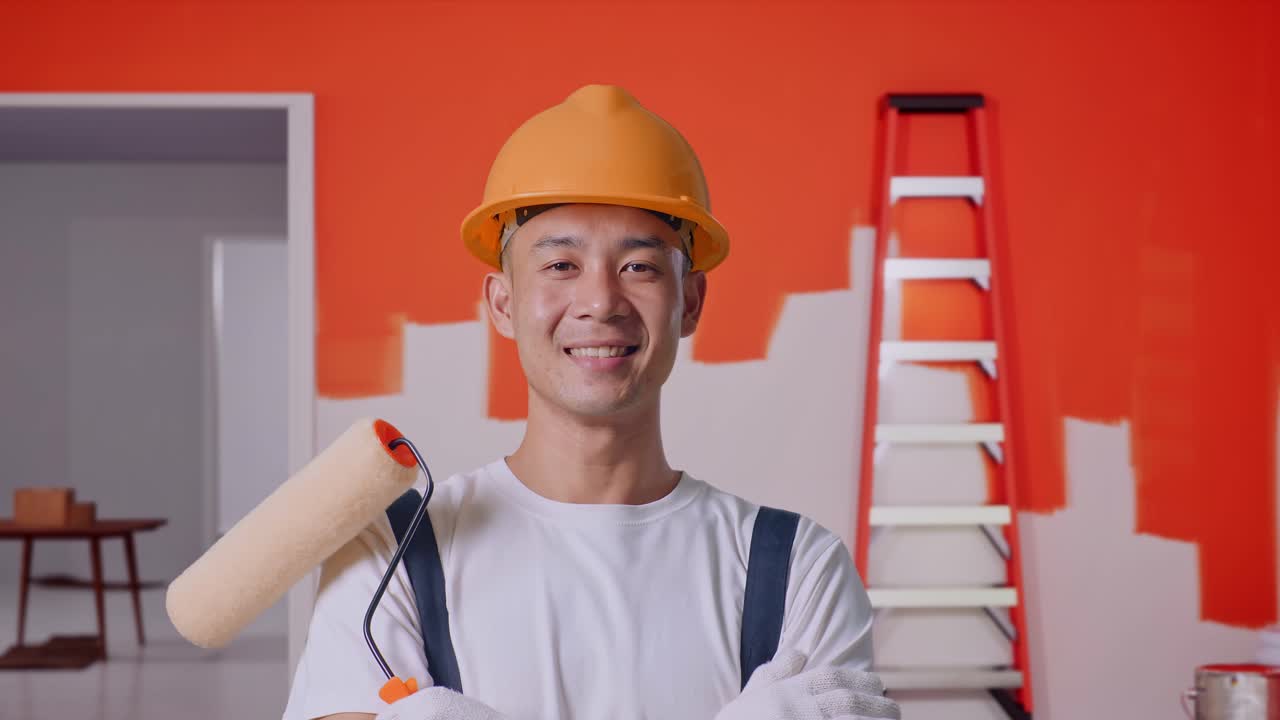 un pintor asiático con casco de seguridad sonriendo y cruzando los brazos mientras está de pie pintando la pared en la habitación