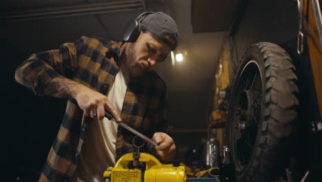 Un-Mecánico-Con-Barba,-Gorra-Y-Auriculares-Trabaja-Con-Una-Lima-En-Un-Banco-De-Trabajo-Y-Escucha-Música-Mientras-Trabaja-En-Un-Taller-De-Estudio.