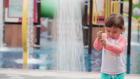 playful little girl at water playground drags rope to start shower waterfall, kid playing refreshing at aqua park on summer day splashing water and get herself wet