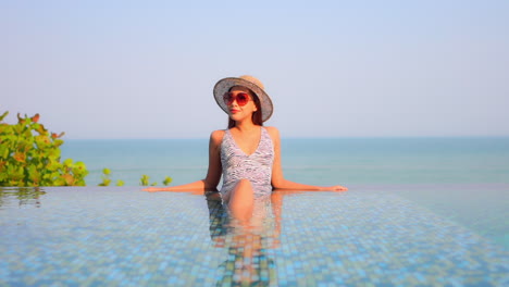 Mujer-Joven-Asiática-Con-Sombrero-De-Sol-Y-Gafas-De-Sol-Rojas-Apoyada-En-El-Borde-Dentro-De-La-Piscina-Infinita-Vista-Frontal-Puesta-De-Sol-En-Un-Cielo-Sin-Nubes,-Espacio-De-Copia-De-Plantilla