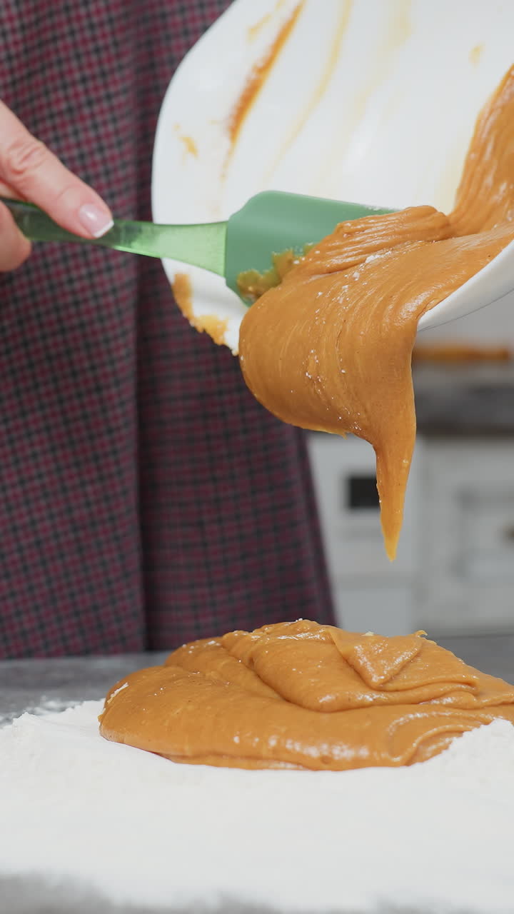 Partial view of woman pouring thick caramel-colored batter into flour on countertop using green spatula, smooth, creamy mixture flows from white bowl