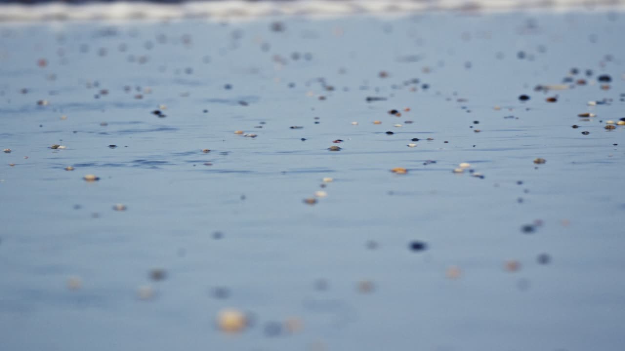 작은 자갈이 있는 해변의 물을 졸졸 흐르는 바람의 낮은 근접 촬영, 뒤이어 밀려오는 파도