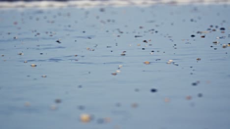 Low-close-up-shot-of-wind-rippling-the-water-of-a-beach-with-small-pebbles,followed-by-a-wave-washing-up