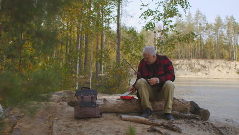 middle-aged man is opening box with fishing baits and lures sitting on log on river shore preparing to angling