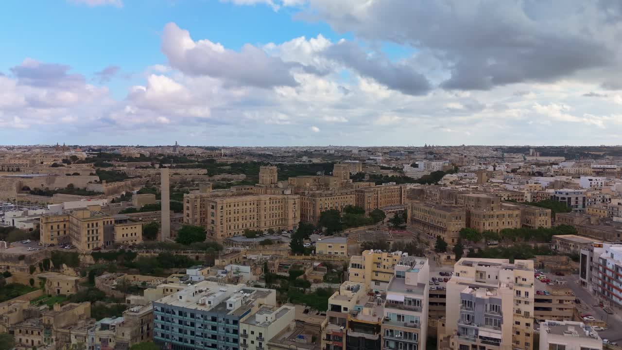 Vista panoramica aerea dello storico complesso dell'Ospedale di San Luca a Pietà, Malta, che mostra i suoi grandi edifici in arenaria, il verde circostante e il paesaggio urbano sotto un cielo blu nuvoloso