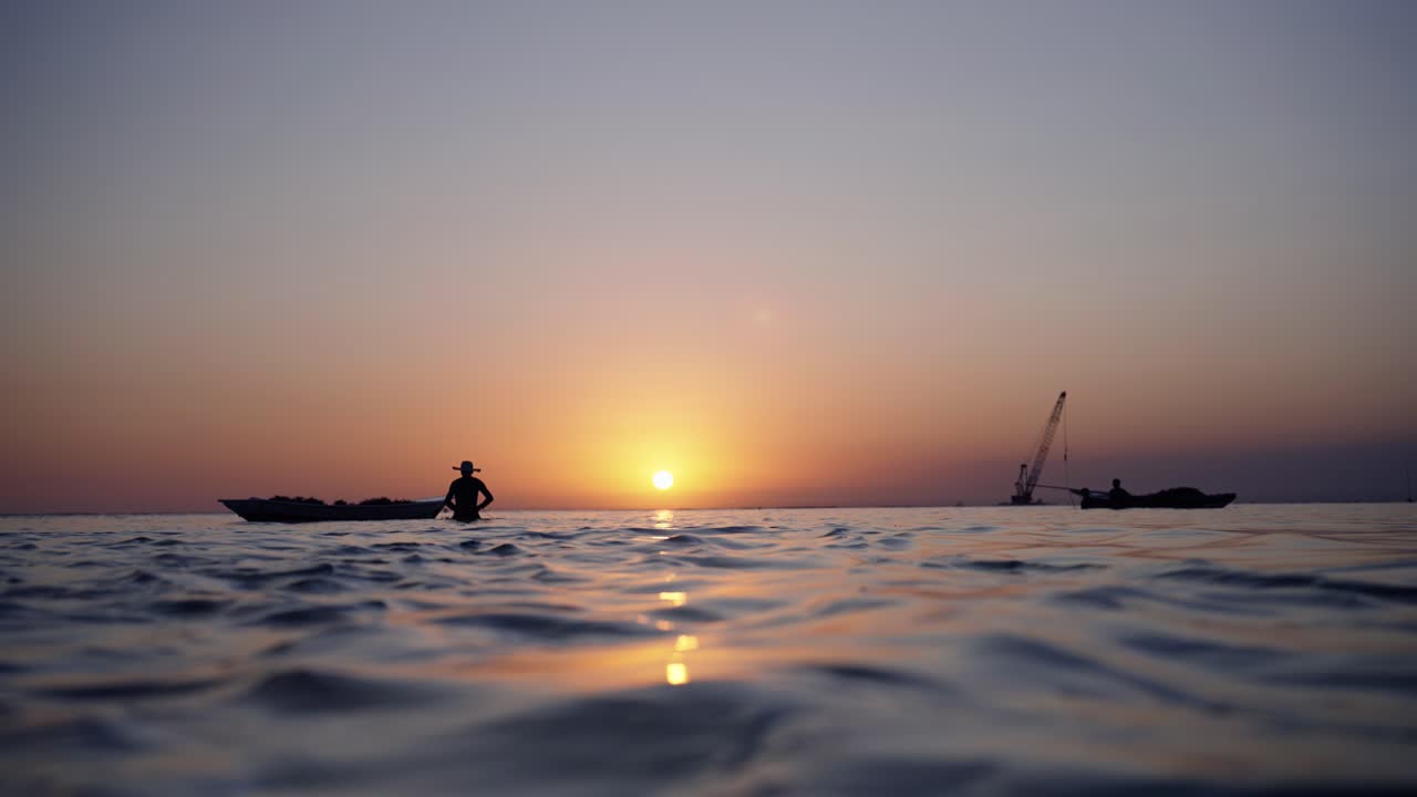 Beautiful sunset clip of a man seaweed farming from his small wooden boat with the golden sunset and a shipwreck in the background