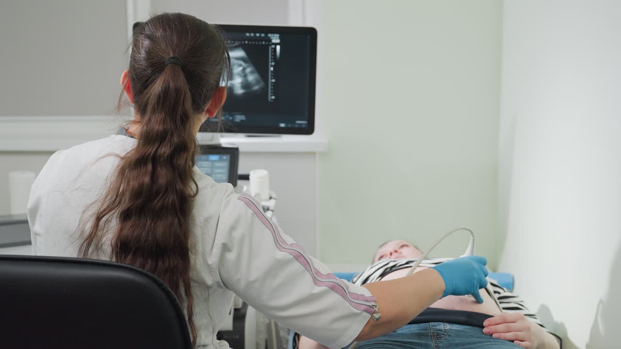Radiology technician conducts abdominal ultrasound on woman lying on clinical bed while focusing closely on monitor displaying real-time imaging data during patient examination inside medical room
