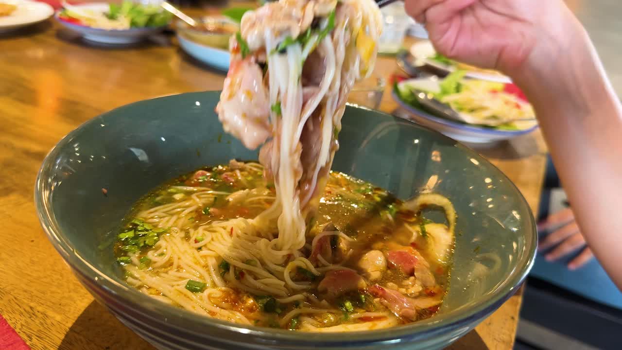 Chopsticks lift rice noodles from a steaming bowl of beef pho, capturing the vibrant dining atmosphere in a Bangkok restaurant