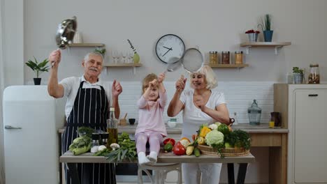 senior woman and man with grandchild girl making a funny dance with strainer and vegetables at home