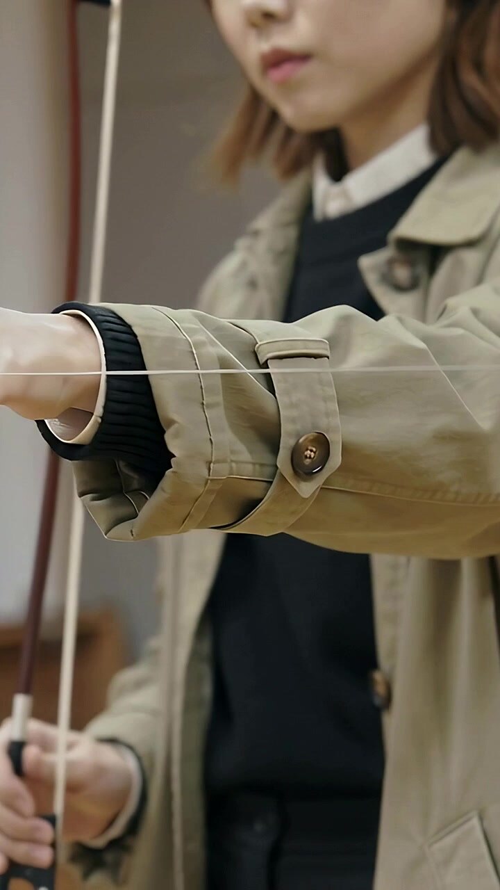 Vertical video: Pinching taut string, woman wearing beige trench drawing bow in studio