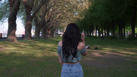 attractive and playful latina woman with black wavy hair walking under the trees in a park in london, looking at the camera and making a follow me gesture, happy with a beautiful smile