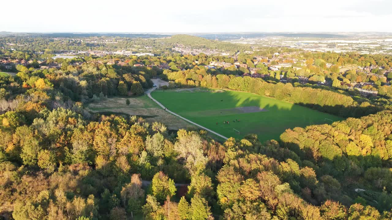 Drone static shot over Parkwood Springs near Sheffield Ski Village, revealing an open field framed by autumn trees, winding paths, and distant homes—where nature meets the edge of the city