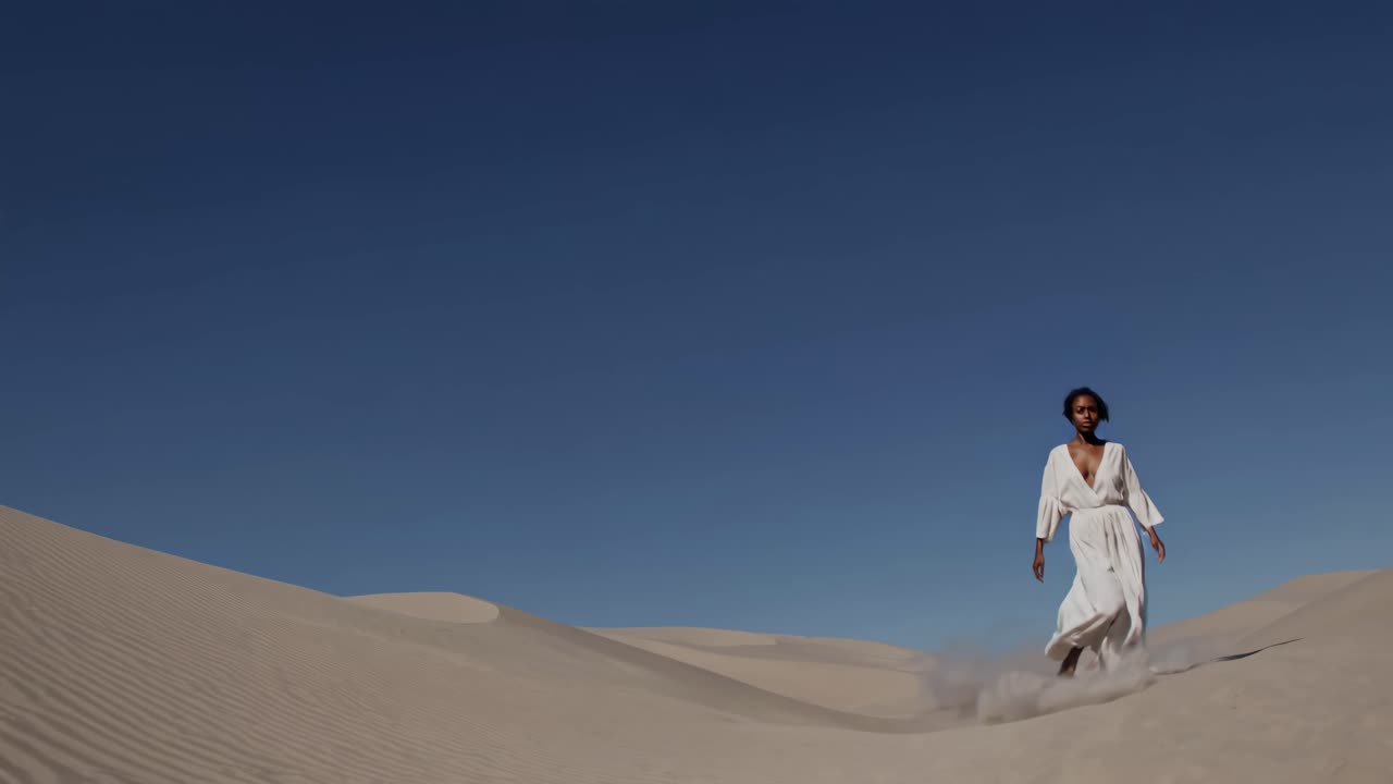 Low-angle video shot of a woman in a flowing white dress walking confidently across sand dunes