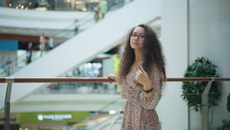 mujer en vestido vintage posa pensativamente coloca la mano en la cintura cerca de la barandilla de hierro, ajustando su cabello en un centro comercial brillante, el borrón de fondo destaca a la gente en la escalera mecánica en movimiento y la planta en maceta cercana