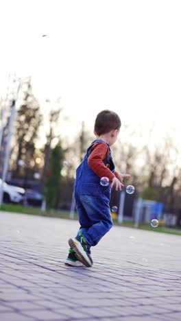 energetic happy caucasian toddler runs to catch soap bubbles. cheerful three-year-old baby having active time outdoors. vertical video.