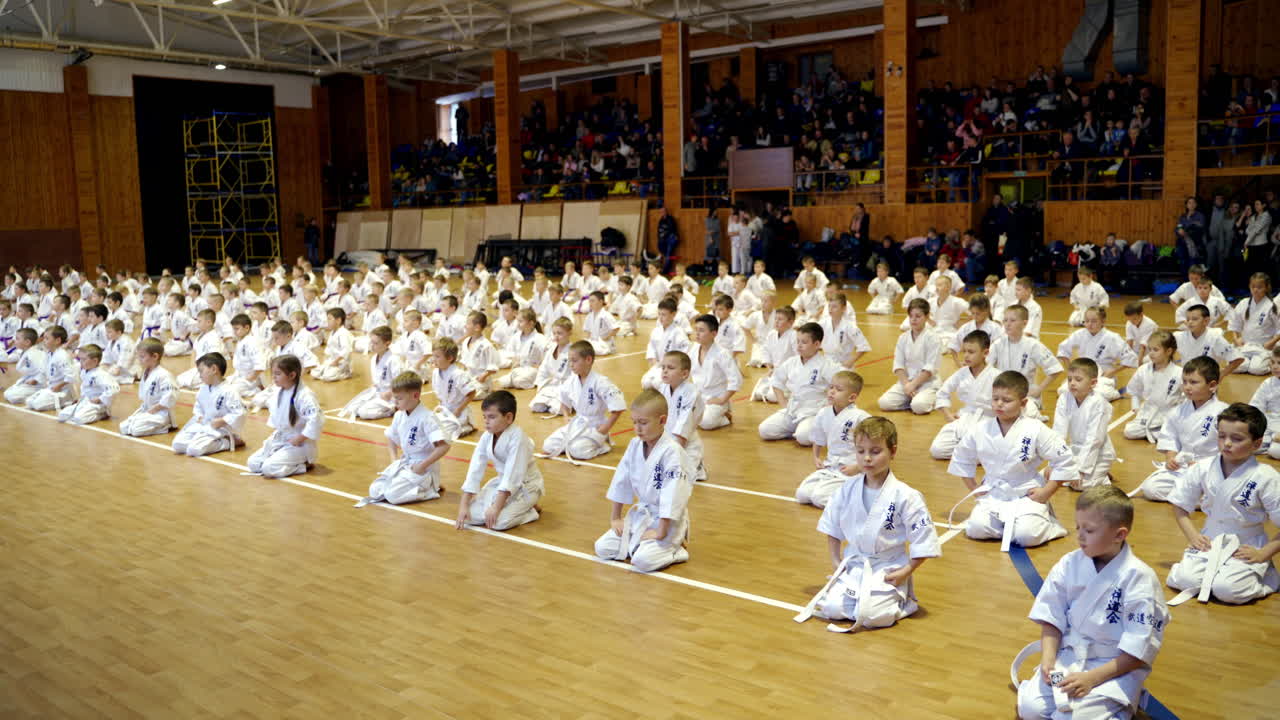 Kids sportsmen in white kimono sit on their knees on the floor. Large group of young karate students sitting with their eyes closed in huge gym.