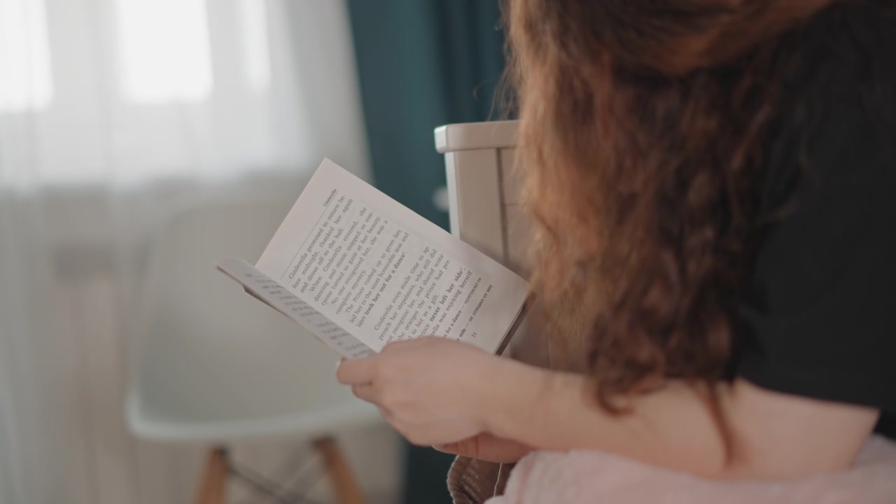 Una donna con i capelli ricci seduta in una stanza illuminata delicatamente legge un quaderno scritto a mano, girando delicatamente le pagine e chinandosi in avanti in una postura concentrata e riflessiva, con un'atmosfera naturale e calma.