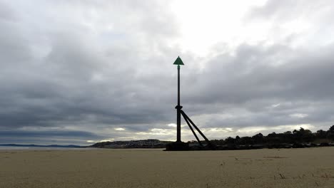 Bewölkte-Gewitterwolken-Im-Zeitraffer,-Die-über-Der-Flutmarkierung-Aus-Metall-An-Der-Küste-Des-Sandstrandes-Vorbeiziehen