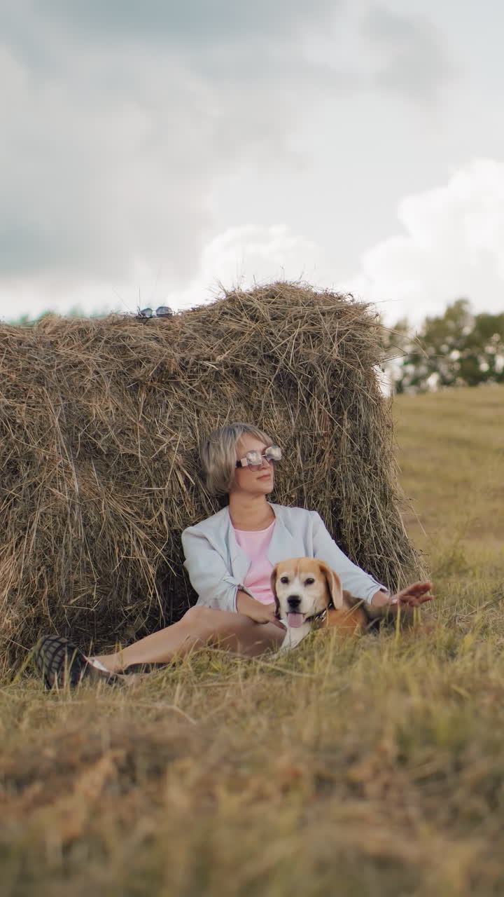 mujer con gafas de sol y traje casual se sienta contra la bala de heno en tierras de cultivo abiertas, acariciando suavemente a su perro, la hierba dorada, las colinas onduladas y el cielo nublado