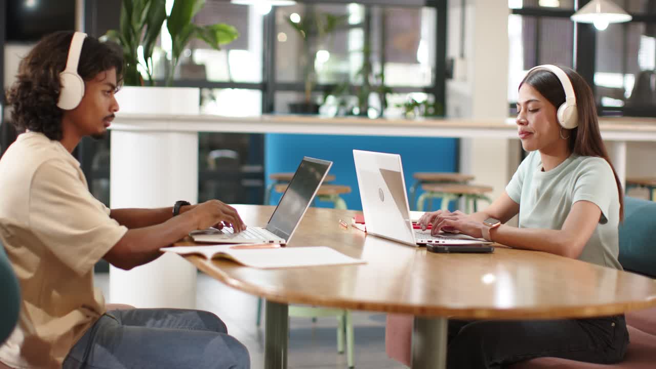 Afroamerikanische Kollegen öffnen Laptops, tippen, lernen mit weißen Kopfhörern an einem Coworking-Tisch