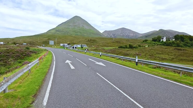 Un veicolo percorre una strada rurale tortuosa attraverso verdi colline e montagne sotto una luce diurna nuvolosa, con un movimento fluido della telecamera e ampie vedute paesaggistiche.